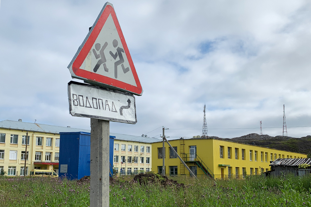 Указатель на водопад недалеко от автобусной остановки в Лодейном. Светло-желтое здание на фоне — действующая сельская школа